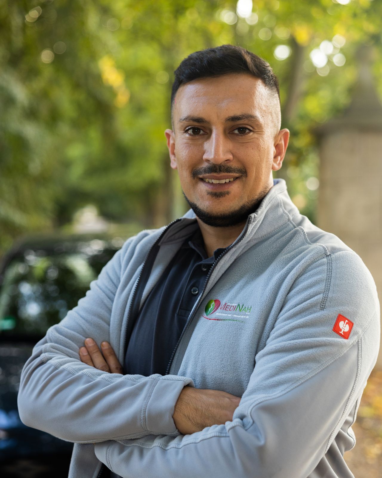 Mitarbeiter des Pflegedienstes MediNah in Dienstjacke, lächelnd vor einem Firmenwagen